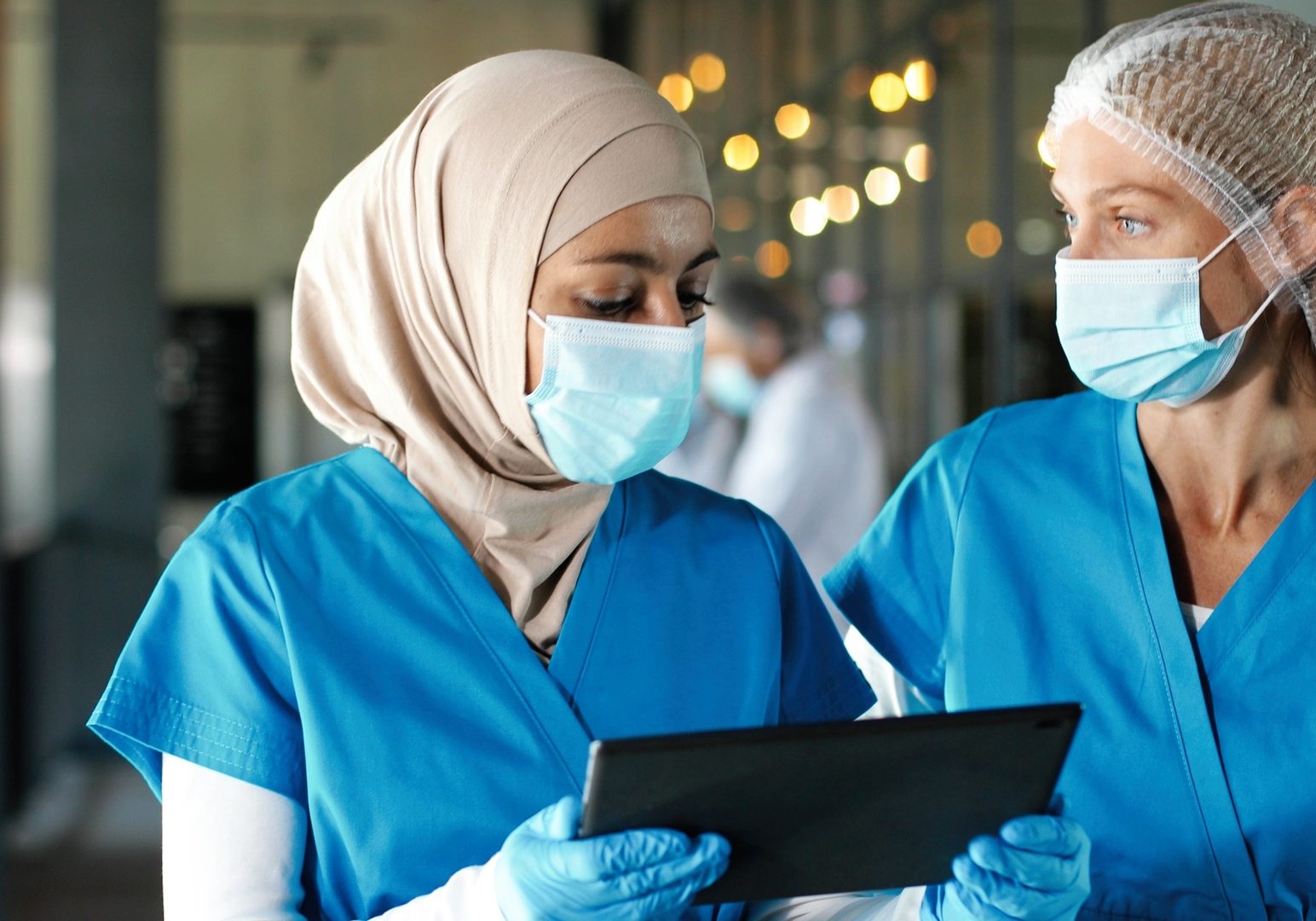 Multi ethnic women doctors in medical masks and gloves walking in hospital and talking about diagnosis. Mixed-races females medics consulting and discussing illness. Covid-19 Nurses with tablet device